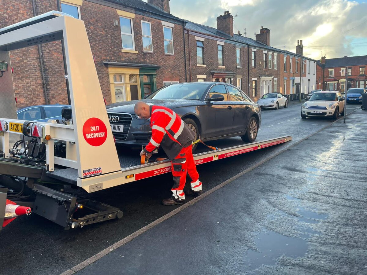Breakdown Man operator in red high-vis crouched at the front wheel of a grey Audi A6 saloon as it loads onto the Breakdown Man recovery truck on a Wigan terraced street