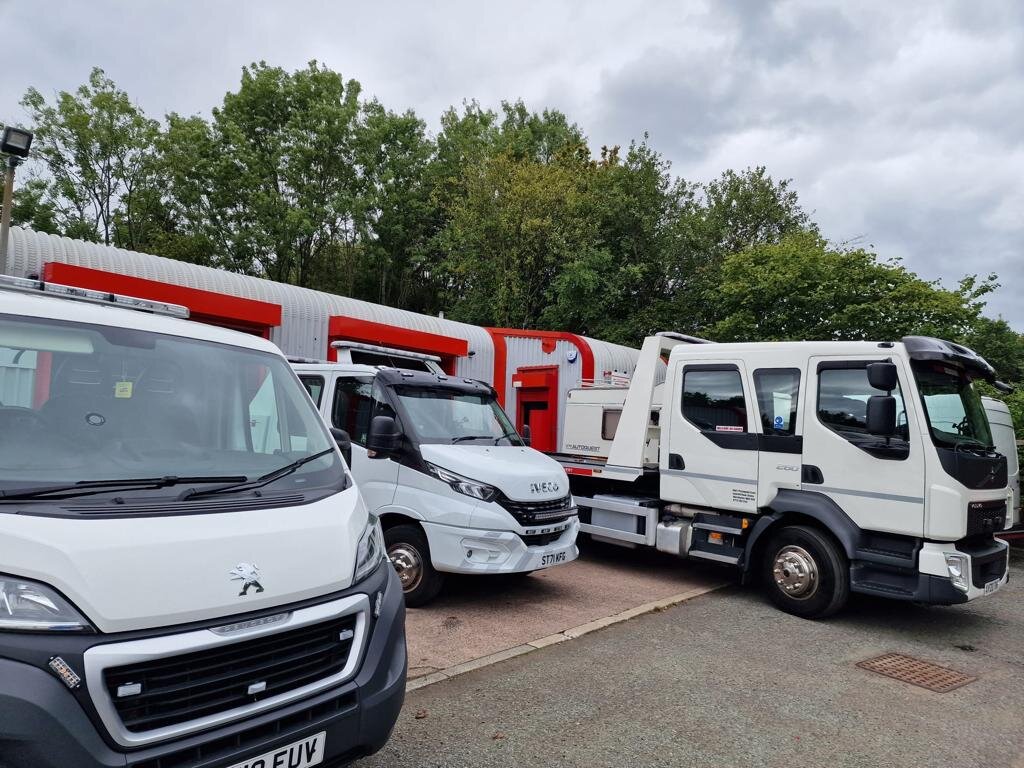 Three Breakdown Man vehicles parked outside the Wigan yard — a white Peugeot Boxer panel van, an Iveco Daily van, and a DAF recovery truck — under cloudy skies.