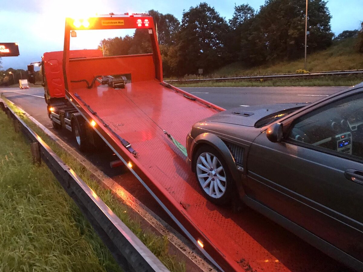 Grey Ford Mondeo loaded onto Breakdown Man's red recovery truck at dusk on the motorway with beacon lights running.