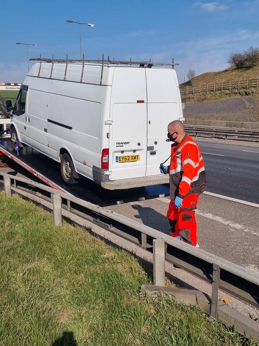 White Ford Transit van loaded onto Breakdown Man's recovery truck beside the motorway, recovery operator in orange hi-vis checking the strap.