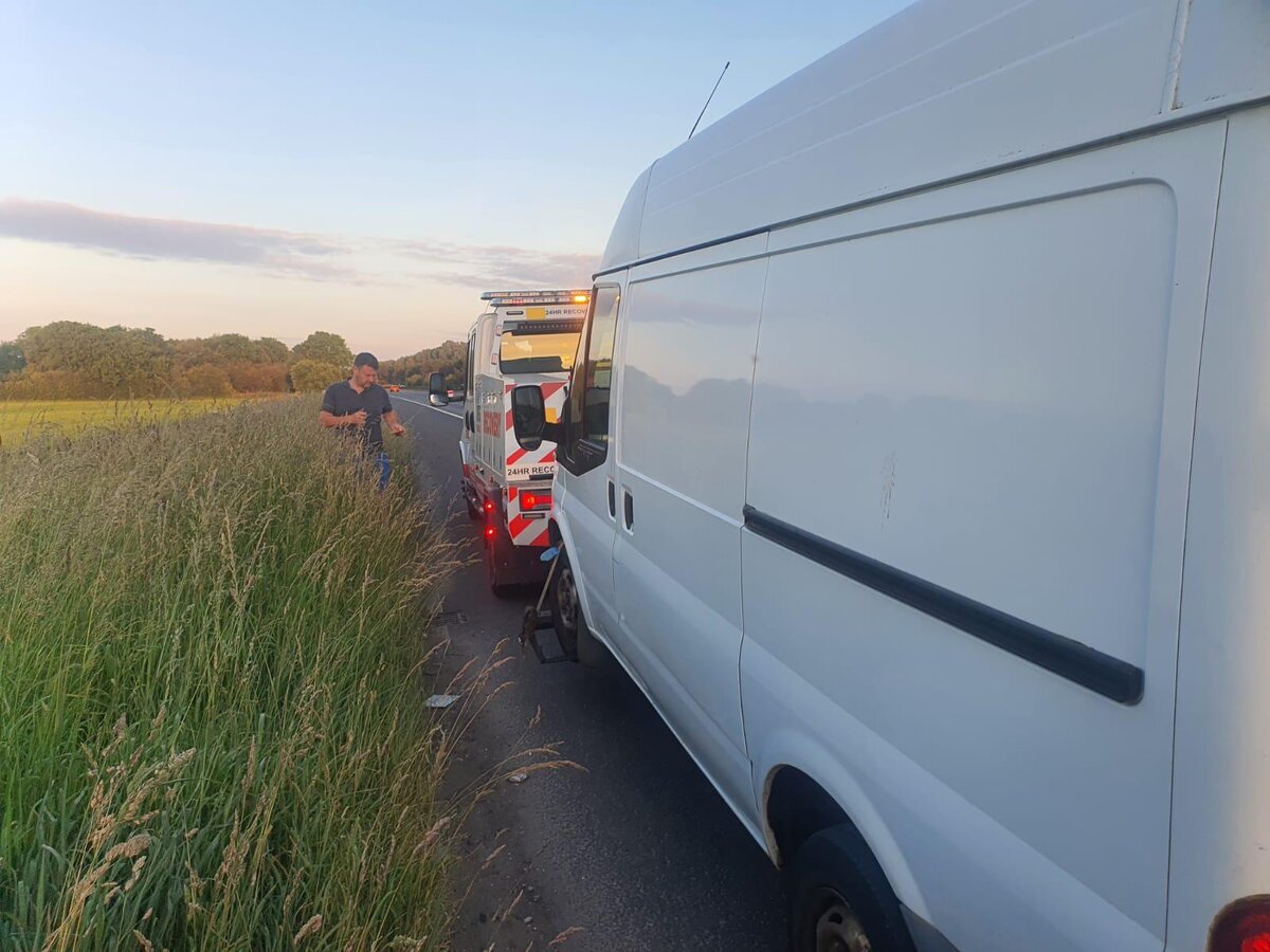 White Ford Transit van pulled onto a rural roadside at dusk with Breakdown Man's recovery truck and hazards behind.
