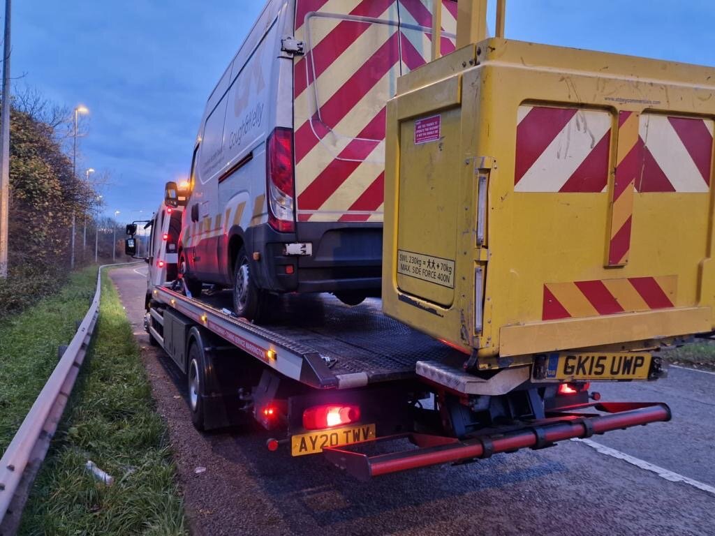 Gough & Kelly silver Iveco van strapped down on Breakdown Man's recovery truck at dusk on the motorway slip road.