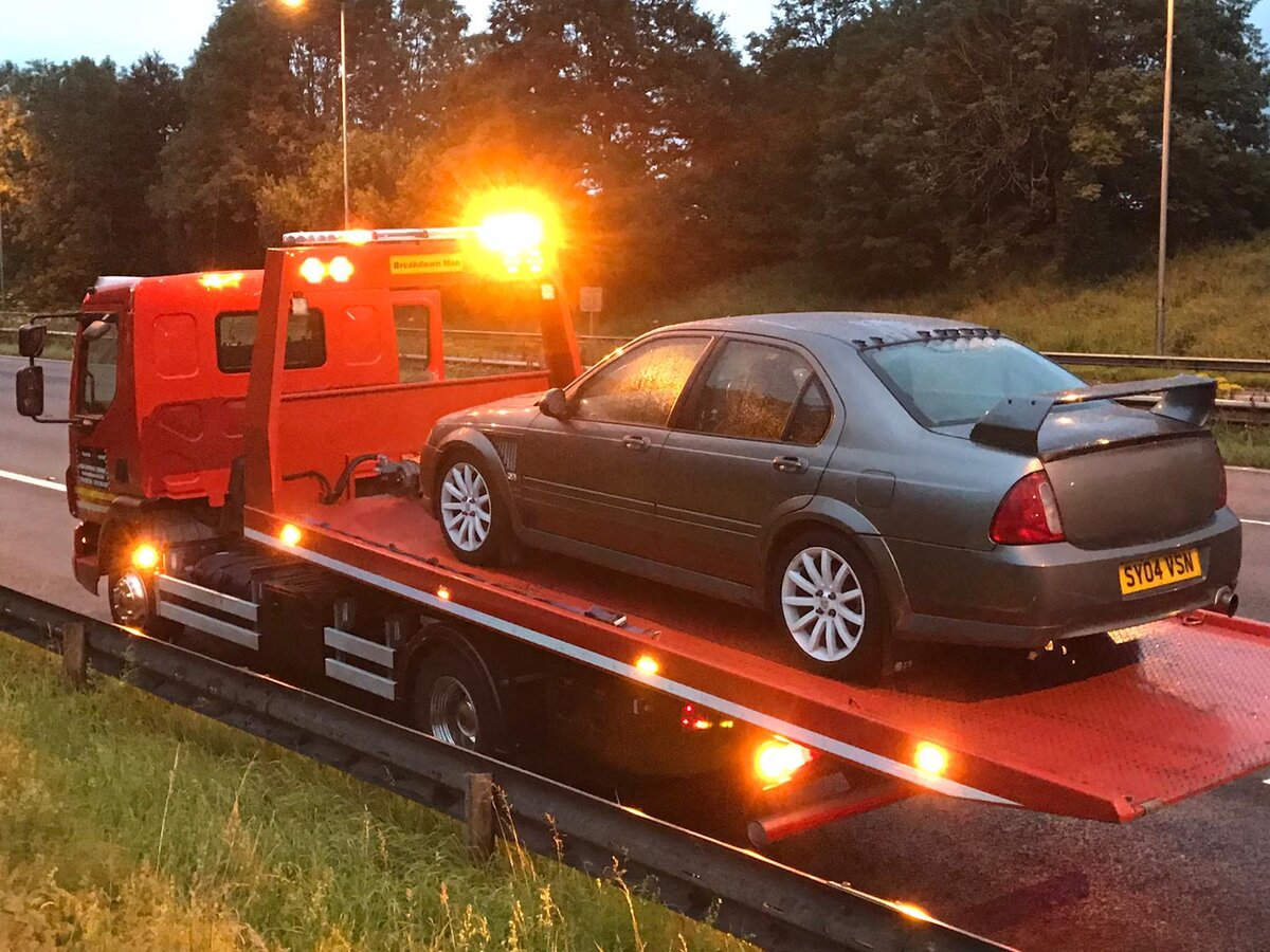 Grey four-door saloon loaded onto a Breakdown Man red recovery truck at dusk with amber beacons lit beside a motorway carriageway.