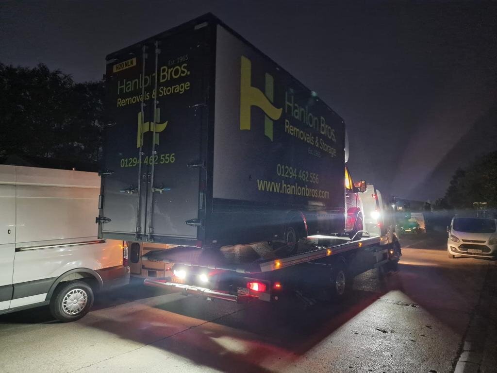 Hanlon Bros Removals & Storage Luton box truck loaded onto a Breakdown Man recovery truck at night, with a white van and other vehicles alongside.
