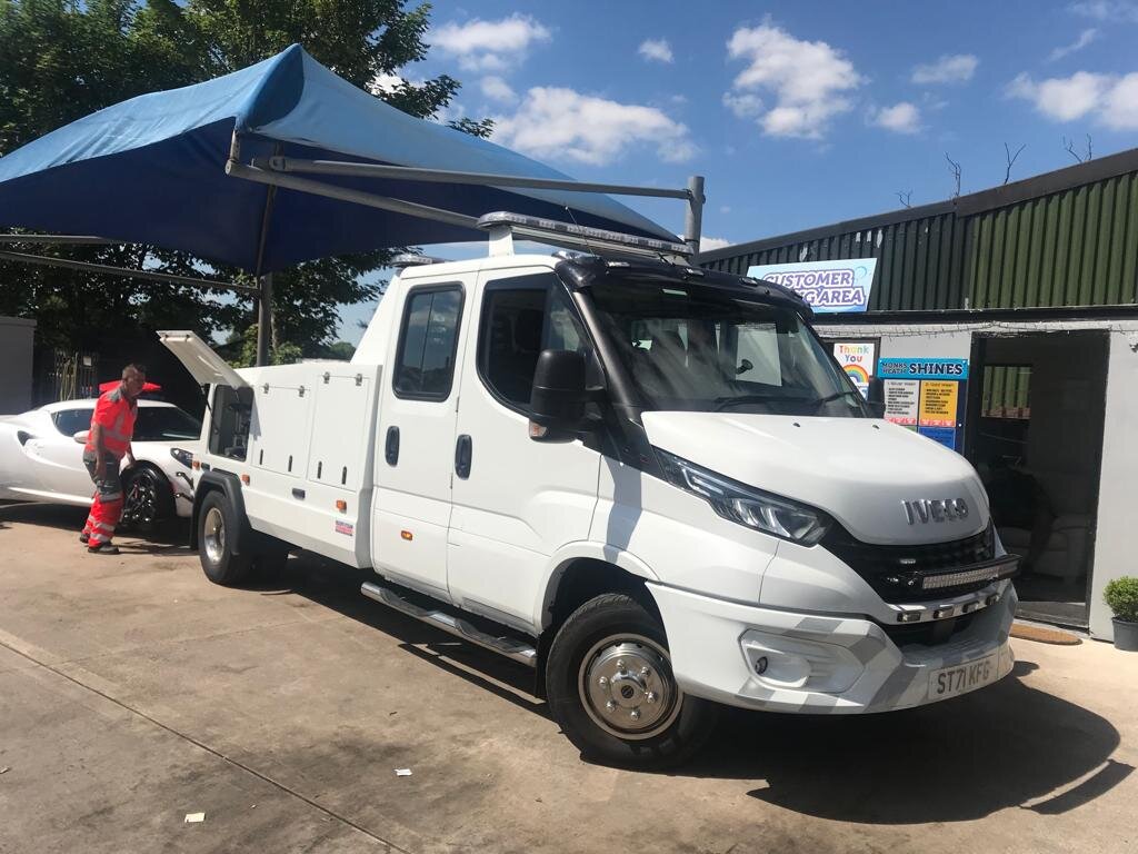 Breakdown Man's white Iveco 24/7 recovery truck parked under the yard canopy with the operator in orange hi-vis and a customer car in the background.