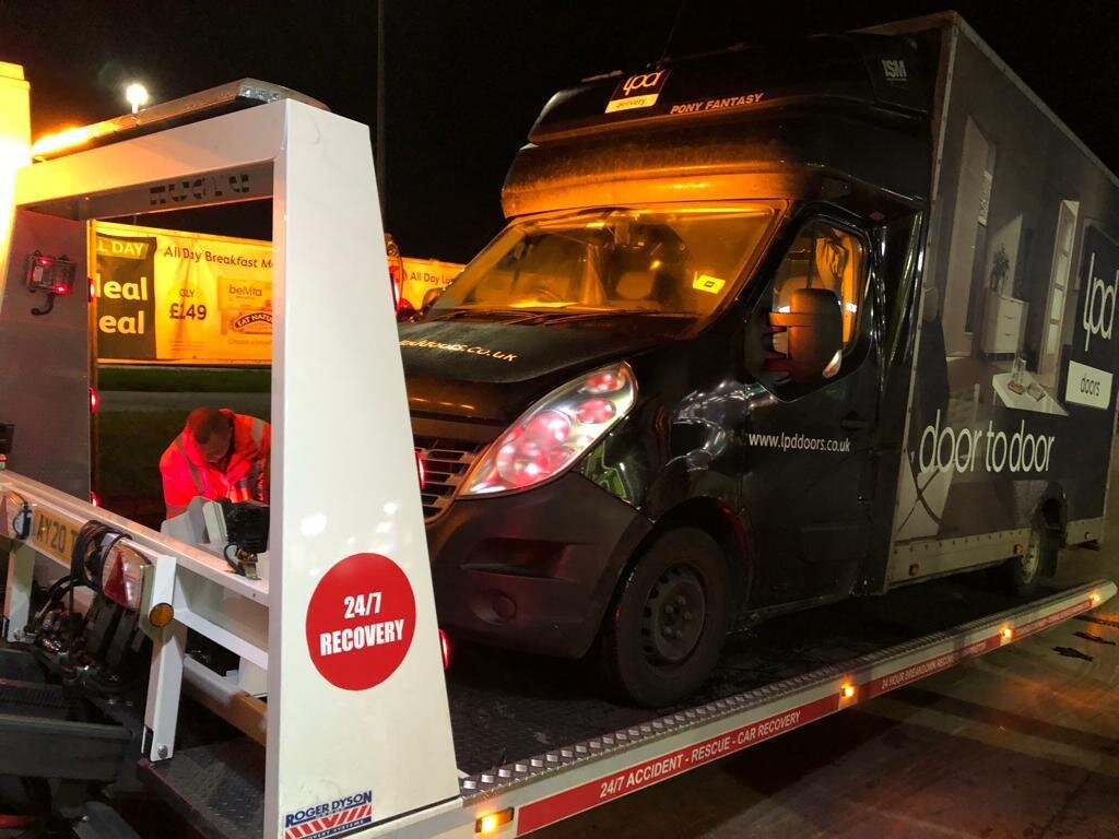 Black LPD Doors-branded Renault Master courier van with side-impact damage being loaded onto a 24/7 Recovery truck at night, recovery operator working alongside.