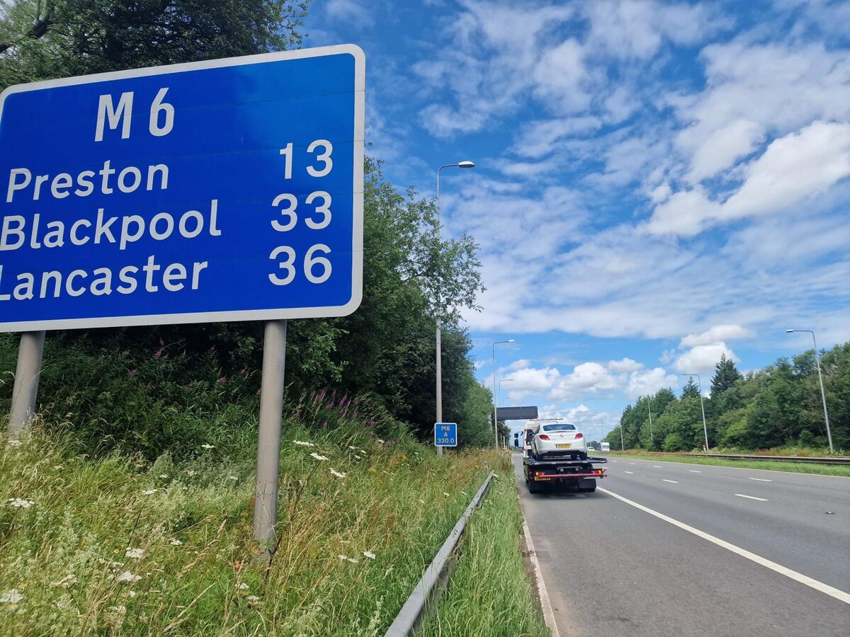 Breakdown Man's recovery truck heading north on the M6 past the Preston, Blackpool and Lancaster mileage sign with a car loaded for recovery.