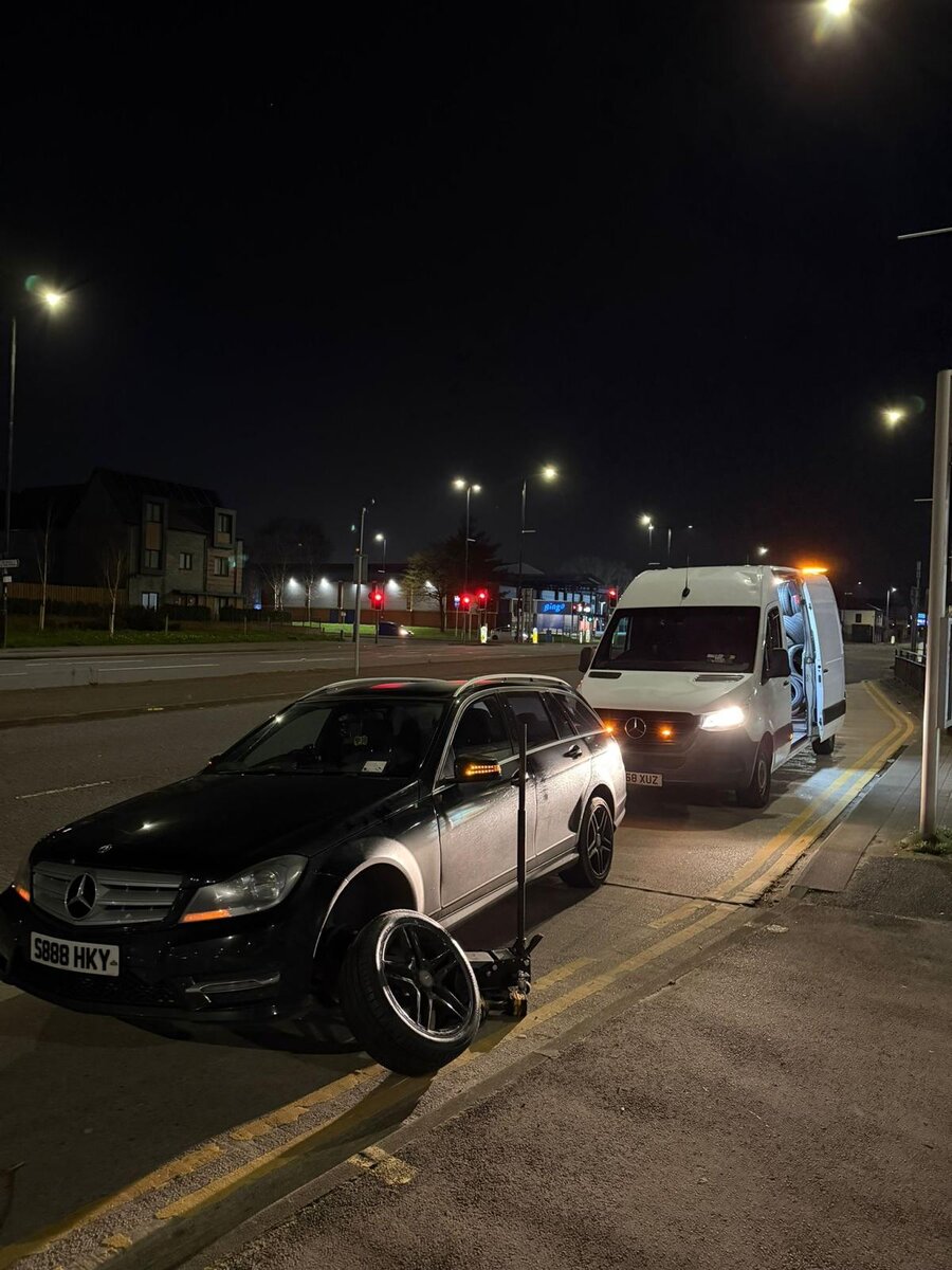 Night-time roadside tyre job — black Mercedes C-Class on a wheel jack at the kerb with the Breakdown Man Mercedes Sprinter mobile tyre van standing by under streetlights.