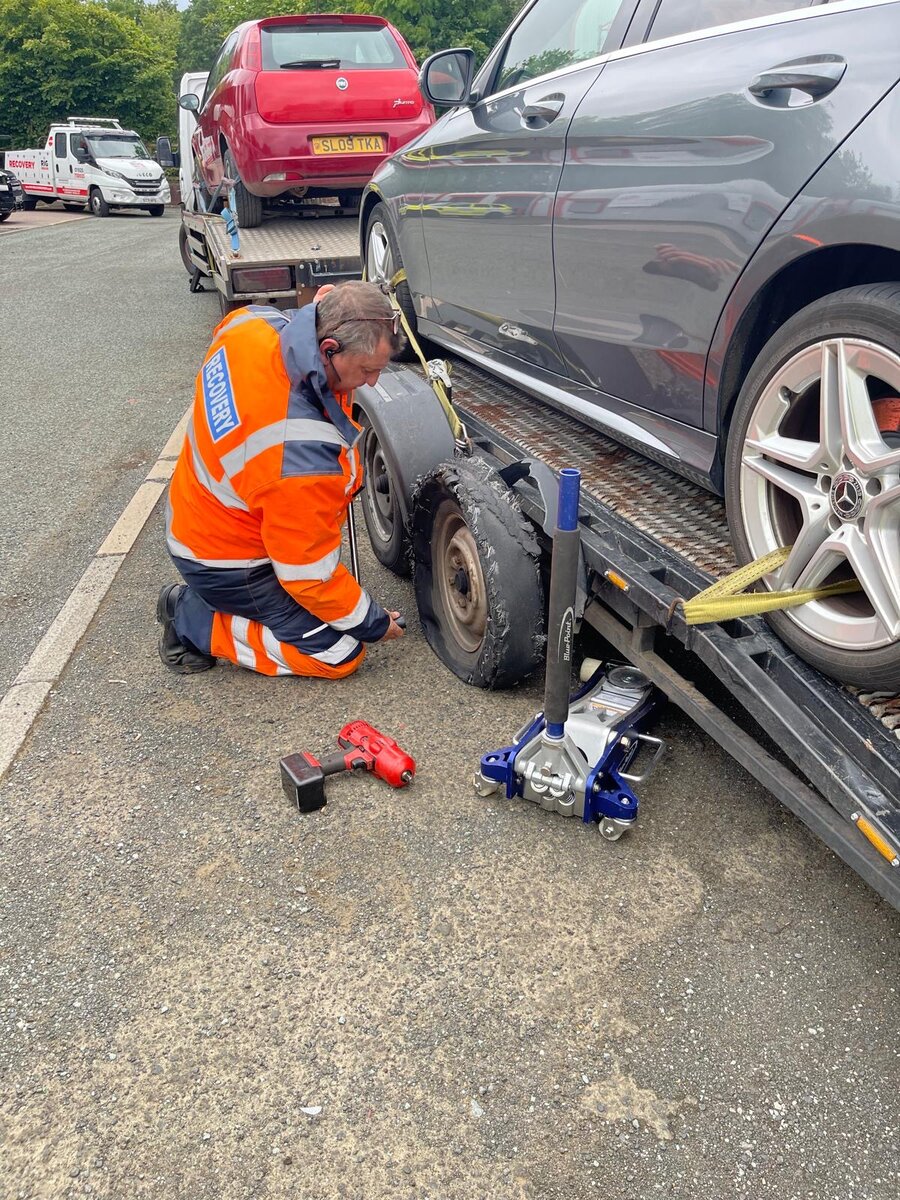 Recovery operator in orange hi-vis strapping a dark Mercedes saloon to the recovery truck with a red trolley jack and recovery dollies.