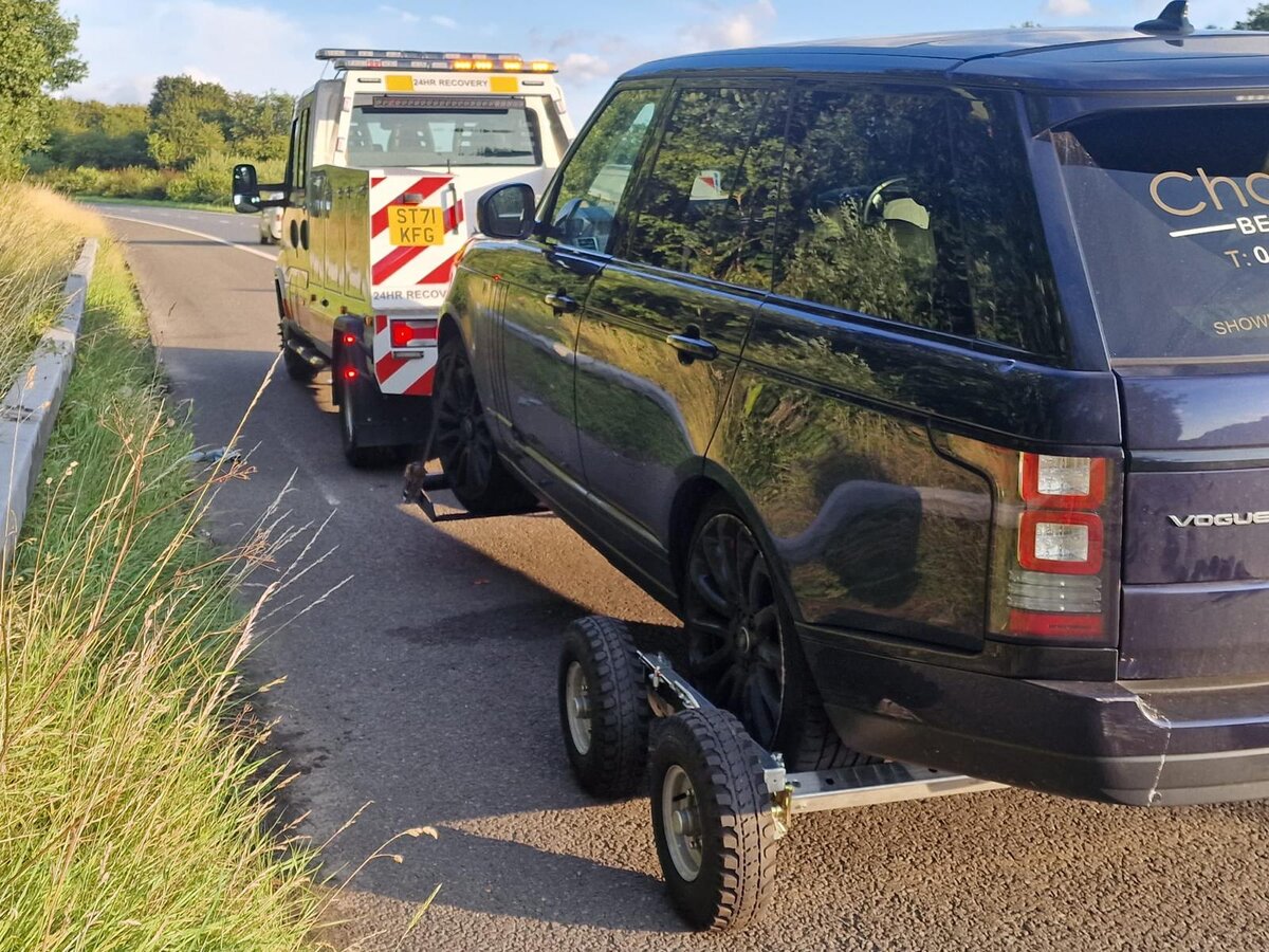 Breakdown Man recovery truck ST71 KFG towing a dark blue Range Rover Vogue on dollies along a country lane.