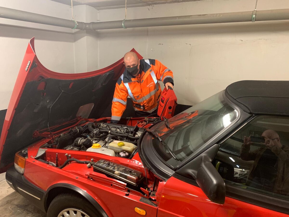 Roadside engine diagnosis on a red Saab convertible — battery booster in hand, bonnet open.