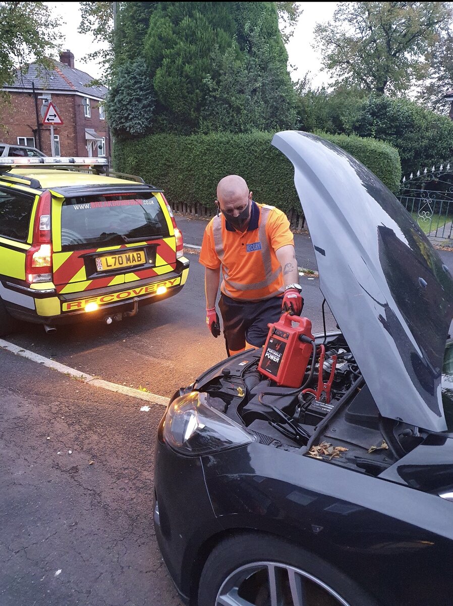 Simon working under the bonnet of a black hatchback at the roadside, Breakdown Man yellow response van parked behind.