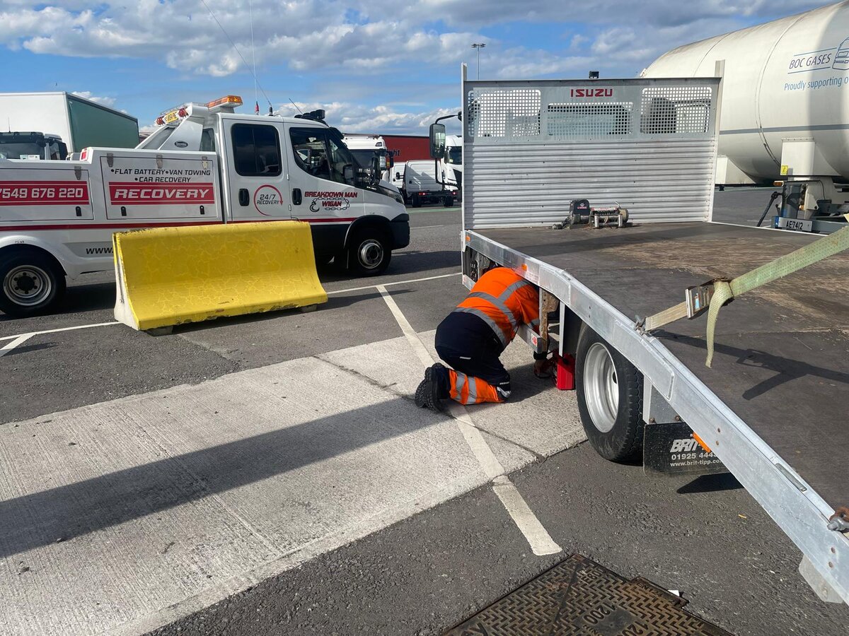 Breakdown Man operator in orange hi-vis kneeling under a recovery truck at the Wigan depot to fit straps before a St Helens job.
