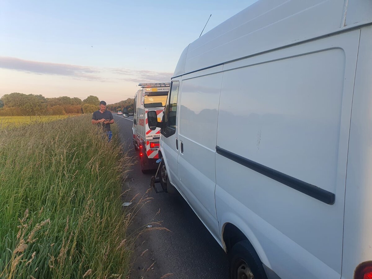 White Ford Transit van pulled over on a rural roadside verge at sunset with a Breakdown Man 24HR Recovery truck behind and the recovery operator stood by the van.