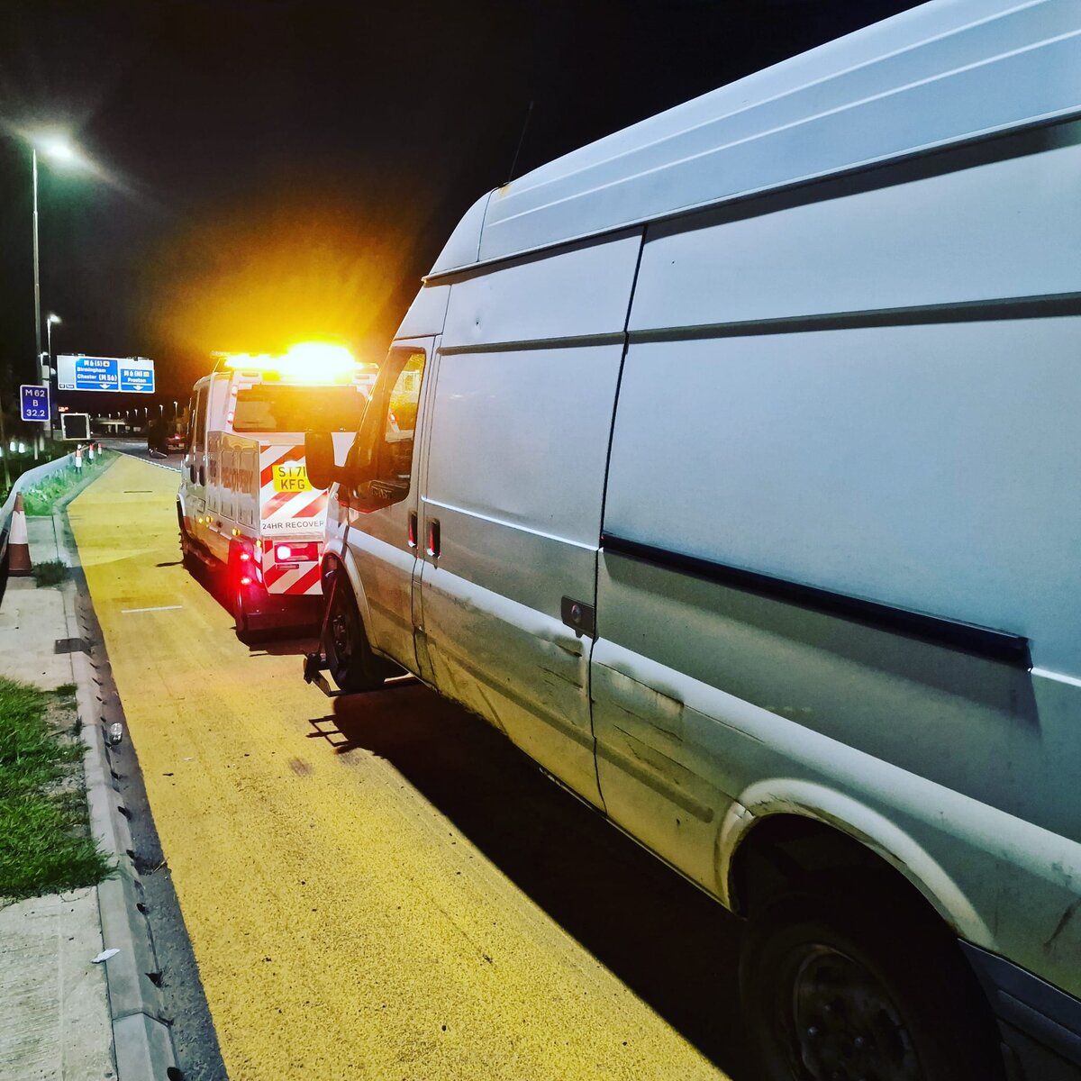 Silver Ford Transit van on the motorway slip road kerb at night with a Breakdown Man 24HR Recovery truck behind, blue motorway sign in the background.