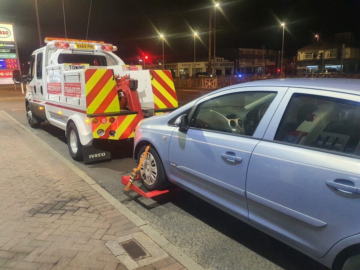 Silver Vauxhall Astra being loaded with wheel-lift recovery dollies onto Breakdown Man's Iveco truck at night near an Esso forecourt.