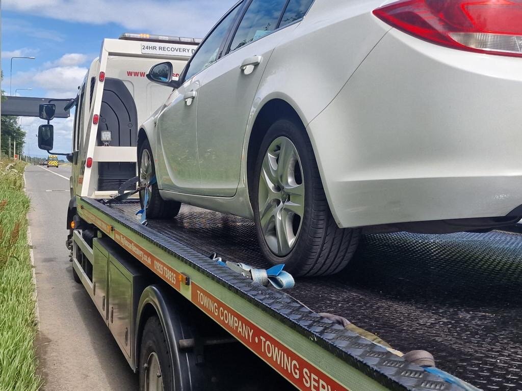 White Vauxhall Insignia saloon strapped down on a Breakdown Man 24HR Recovery truck on a rural roadside verge.