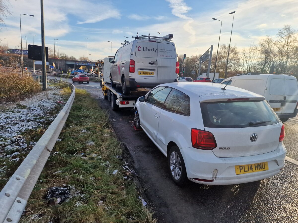 White Volkswagen Polo parked on a frost-dusted roadside verge alongside a Liberty-branded service van and a Breakdown Man recovery truck on the slip road.