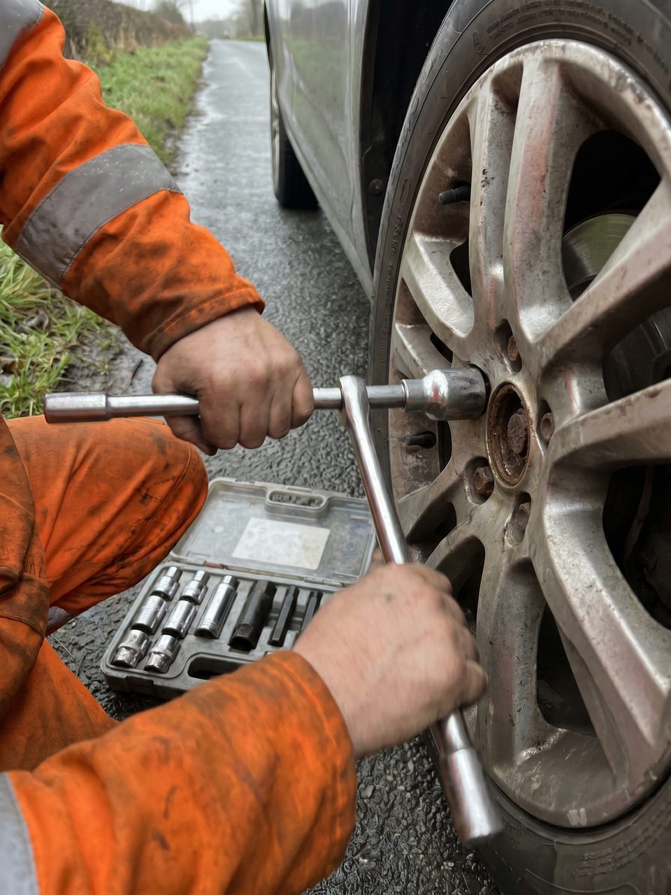 A stubborn locking wheel nut being removed from a car's alloy wheel with a specialist extraction socket, key sockets laid out at the roadside.
