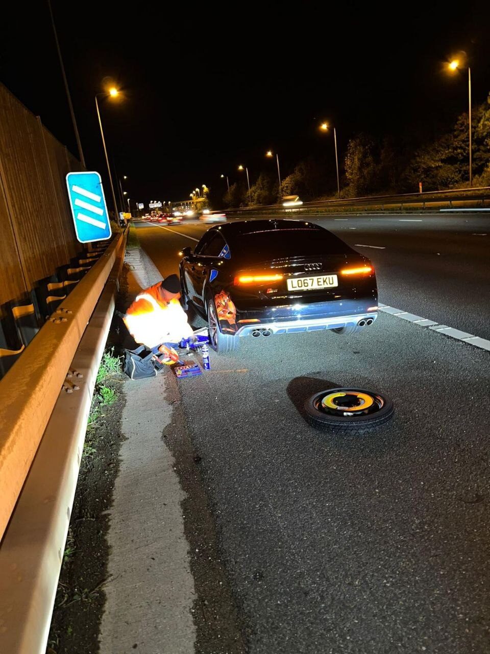 Breakdown Man changing a wheel at the roadside at night, hi-vis on and space-saver spare ready.