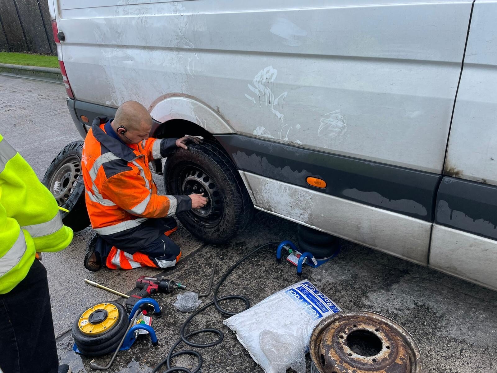 Breakdown Man changing a van wheel kerbside in daylight with tools laid out.